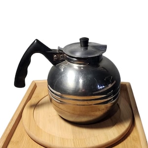 May include: A silver coffee pot with a black handle and a lid. The pot has a shiny finish and is sitting on a wooden surface.