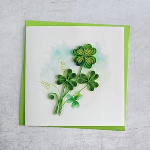 Könnte beinhalten: Handgefertigte Grußkarte mit einem Quilling-Kleeblatt-Design. Die Karte hat einen weißen Hintergrund mit einer Aquarellwaschung und einem grünen Rand. Die Kleeblätter sind in verschiedenen Grüntönen gehalten.
