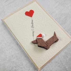 May include: A Christmas card with a brown origami dog wearing a Santa hat, holding a red heart balloon. The words "merry Christmas" are written in cursive. The card is on a cream-coloured background.