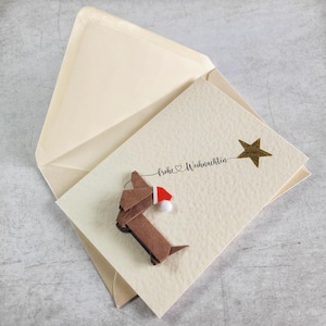 May include: A cream Christmas card featuring a brown origami dachshund wearing a Santa hat. The card has the words "Frohe Weihnachten" and a gold star. The card is inside a cream envelope.
