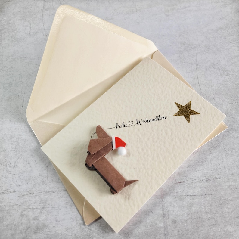 May include: A cream Christmas card featuring a brown origami dachshund wearing a Santa hat. The card has the words "Frohe Weihnachten" and a gold star. The card is inside a cream envelope.