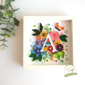 Può includere: Un'opera d'arte in carta incorniciata in bianco con una lettera A bianca circondata da fiori di carta colorati, foglie e una farfalla. Lo sfondo è bianco con piccoli cerchi.