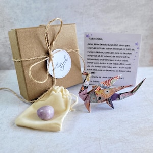 Op de afbeelding: Een cadeau set met een bruine doos vastgebonden met touw, een kleine beige zak met een paarse steen en een kleurrijke origami draak. Een kaart met Duitse tekst is ook inbegrepen, met de woorden "Liebe Emilia" zichtbaar.
