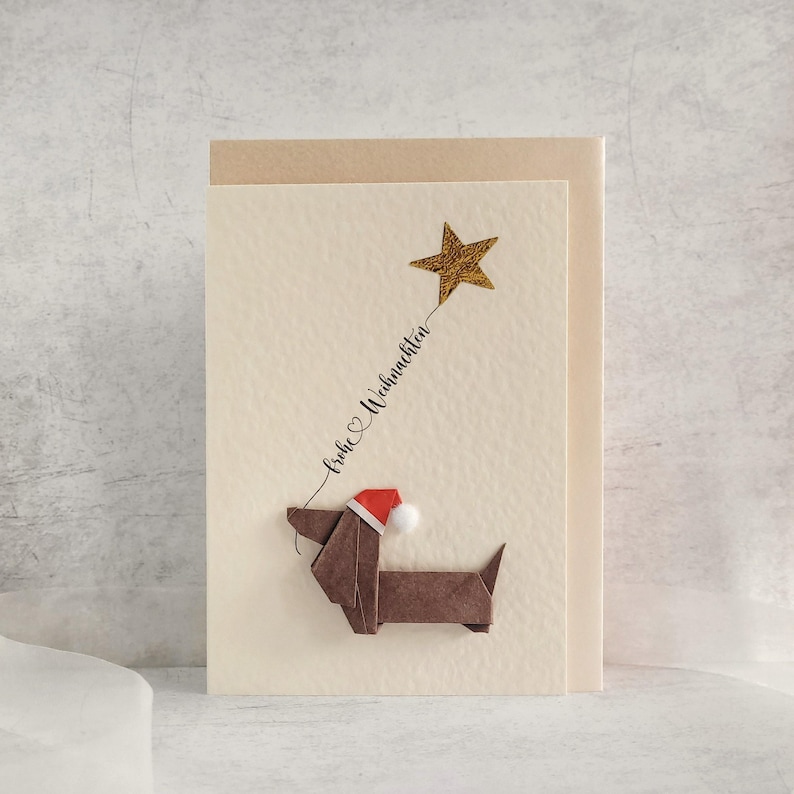 May include: A Christmas card with a brown origami dachshund wearing a Santa hat. A gold star is connected to the dog with cursive text that reads "Frohe Weihnachten". The card has a cream background and a tan envelope.