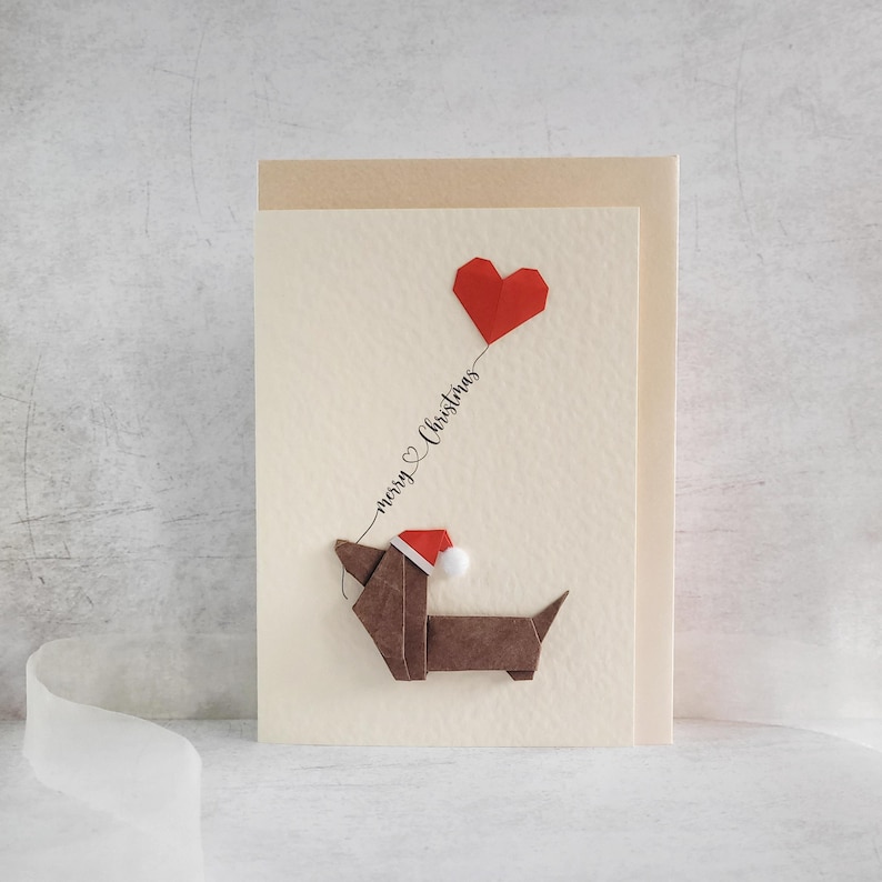 May include: A Christmas card with a brown origami dachshund wearing a Santa hat, connected to a red heart-shaped balloon by a string. The words "merry Christmas" are written on the string. The card has a cream background.