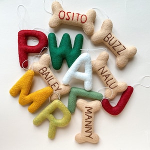 May include: A set of felt dog bone ornaments with names embroidered on them. The names are: Osito, Buzz, Bailey, Nalu, Manny, A, W, J, M, and P.