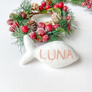 May include: White felt fish ornament with the name "LUNA" embroidered in pink thread. The ornament is hanging from a green pine wreath decorated with red berries and faux snow.