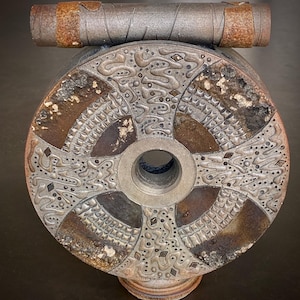 May include: A round, silver and brown metal sculpture with a circular design. The sculpture has a hole in the center and is mounted on a smaller, round base.