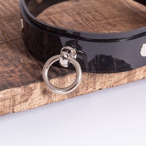 May include: A black leather choker with a silver ring and two silver studs. The choker is on a wooden surface.