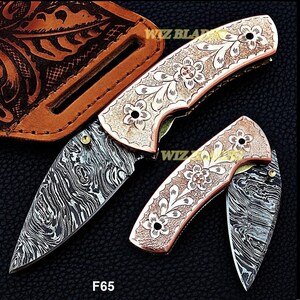 Puede incluir: Dos navajas con diseños florales y hojas de acero de Damasco. Los mangos tienen un acabado color cobre con patrones florales grabados. Una funda de cuero marrón es visible en el fondo. Los textos "WIZ BLADES" y "F65" son visibles.