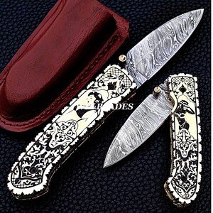 May include: Two ornate folding knives with Damascus steel blades. The handles feature intricate carvings and a cream-colored inlay with a woman's silhouette. A burgundy leather sheath is also present. The blades are open and closed.