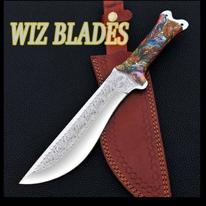 May include: A silver knife with a decorative etched blade and a colorful, multi-colored, patterned handle. The knife is in a brown leather sheath with a gold button. The text "WIZ BLADES" is visible in the image.