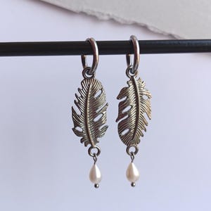 Può includere: Un paio di orecchini a forma di piuma color argento con piccole gocce di perle bianche a forma di goccia. Gli orecchini sono sospesi da cerchietti argentati e sono esposti su sfondo bianco.