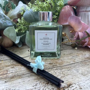 Puede incluir: Una botella de vidrio transparente con una tapa plateada y una etiqueta blanca que dice "Aquamarine Reed Diffuser Jasmine and Geranium 100ml". La botella está sobre una superficie de madera con difusores de caña negros y una cinta azul claro.