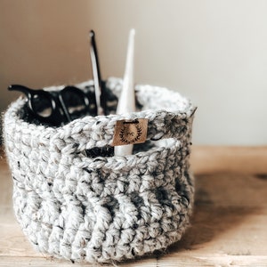 Puede incluir: Una cesta de crochet gris con una etiqueta de cuero marrón que dice "RYC" con un diseño de corona. La cesta está llena de pinzas para el pelo negras y un objeto blanco.