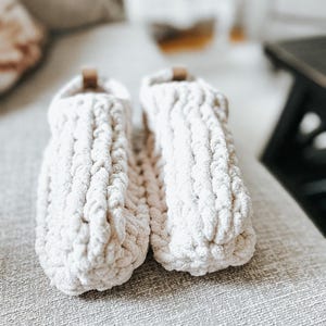 Peut inclure: Une paire de chaussons en tricot épais de couleur crème. Les chaussons ont un aspect épais et texturé et un petit accent en cuir marron sur le dessus. Ils sont conçus pour être portés à l'intérieur et sont posés sur une surface aux tons neutres.