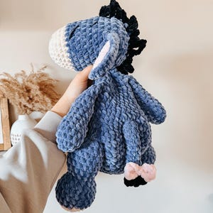 May include: A plush, blue donkey stuffed animal with a white muzzle and pink inner ears. The donkey has a black mane and a small pink bow on its tail. The toy is made of a soft, textured material.