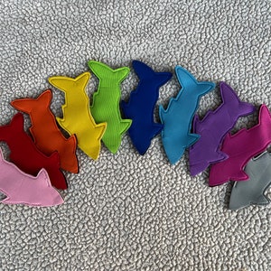 Puede incluir: Un conjunto de diez formas de tiburón de fieltro en colores arcoíris. Los tiburones están dispuestos en semicírculo, con el tiburón rosa en la parte inferior y el tiburón gris en la parte superior.