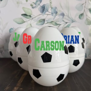 May include: Four white plastic Easter eggs decorated with a black and white soccer ball design. Each egg has a name written on it: "J", "GR", "CARSON", and "BRIAN".