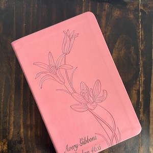 May include: A pink journal with a floral line drawing of lilies. The journal has the text "Avery Gibbons Psalms 46:5" in a script font. The journal is closed and resting on a dark wooden surface.