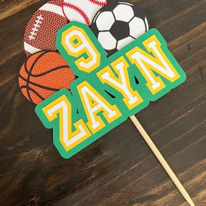 Könnte beinhalten: Ein grüner und gelber Sport-Kuchentopper mit dem Namen "ZAYN" und der Nummer "9" darauf. Der Topper zeigt einen Basketball, einen American Football, einen Baseball und einen Fußball.