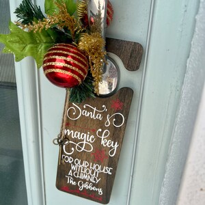 Könnte beinhalten: Ein hölzerner Türkranz mit dem Text "Santa's magic key for our house without a chimney The Gibbons" und einem kleinen Schlüsselanhänger. Der Kranz ist mit einem rot-goldenen, gestreiften Ornament und grün-goldenem Laub verziert.
