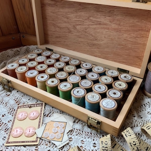 May include: A wooden storage box containing thirty-five spools of thread in various colours, including shades of blue, green, orange, and grey. Two cards of light pink and white buttons are present, along with a yellow measuring tape.