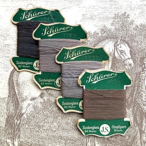 May include: A collection of vintage thread spools in various shades of gray and brown. Each spool has a green label with the name "Schürer" and the text "Seidenglanz 20 Meter". The background features a sepia-toned illustration of a horse.