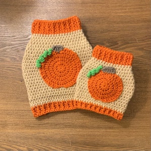May include: Two crocheted cozies, one larger than the other, in a beige color with orange trim. Each cozy features a large, orange pumpkin applique with green leaves. The items are likely designed for fall or Halloween decor.