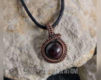 May include: A red garnet gemstone pendant wrapped in copper wire, hanging from a black cord. Garnet - January Birthstone.