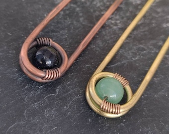 May include: Two hair sticks with wire wrapped around the ends. One stick is copper with a black stone bead and the other is gold with a green stone bead.