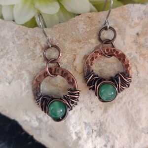 May include: A pair of copper earrings with a hammered circle design and a green stone in the centre. The earrings are made with wire wrapped around the stone and circle.