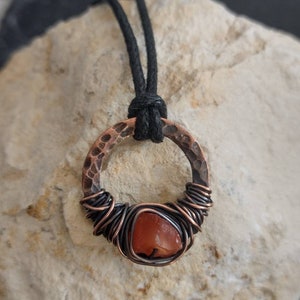 May include: A copper wire wrapped carnelian gemstone pendant with a hammered copper circle. The pendant is on a black cord.