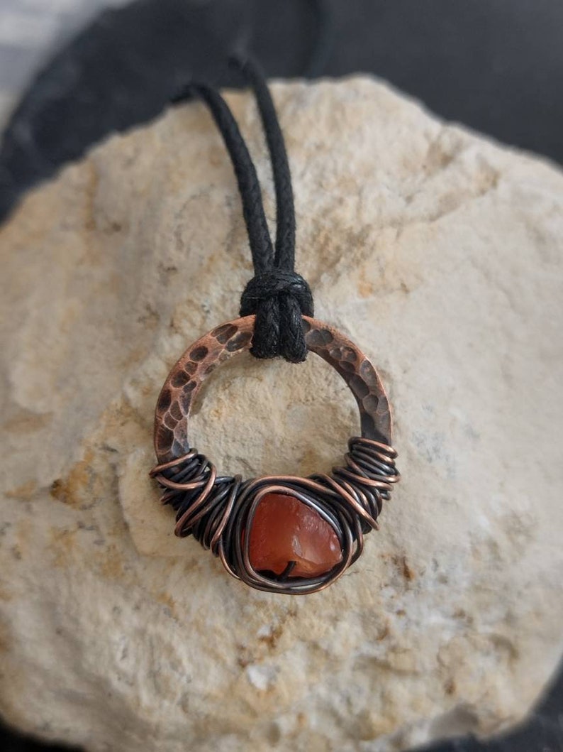May include: A copper wire wrapped carnelian gemstone pendant with a hammered copper circle. The pendant is on a black cord.
