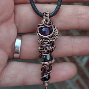 Op de afbeelding: Een hanger gewikkeld met koperdraad met een amethist, hematiet, rookkwarts en een zwarte onyx steen. De hanger is bevestigd aan een zwart koord.