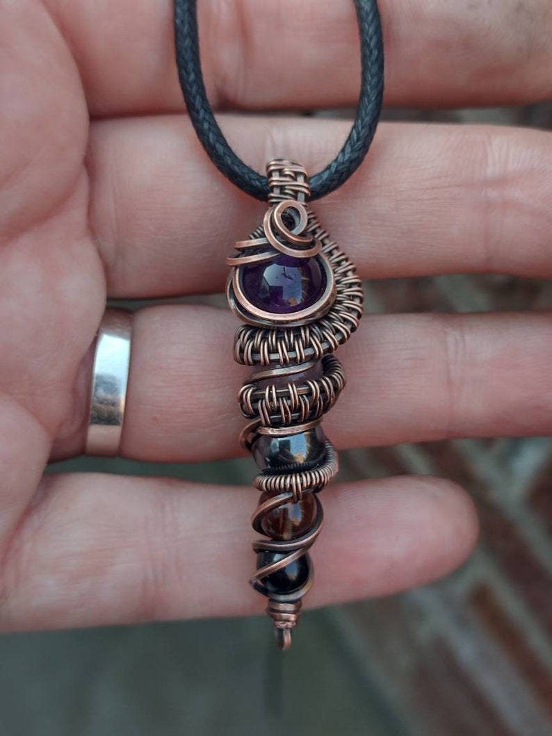 Op de afbeelding: Een hanger gewikkeld met koperdraad met een amethist, hematiet, rookkwarts en een zwarte onyx steen. De hanger is bevestigd aan een zwart koord.