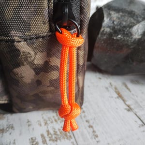 May include: A camouflage patterned fabric bag featuring a black zipper closure. A bright orange paracord pull is attached to the black zipper slider, tied with a knot at the top and a larger knot at the bottom to provide a secure grip.