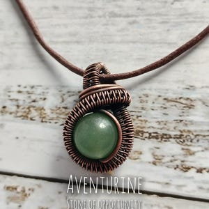 May include: A round green aventurine gemstone pendant wrapped in copper wire, suspended on a brown leather cord. The text below the pendant reads AVENTURINE STONE OF OPPORTUNITY. The wire wrapping features a coiled design around the stone.