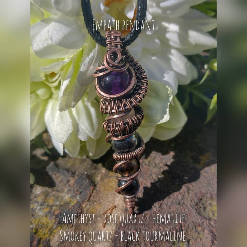 Op de afbeelding: Hanger omwikkeld met koperdraad met amethist, rozenkwarts, hematiet, rookkwarts en zwarte toermalijn stenen. "Empath Pendant" staat boven de hanger.