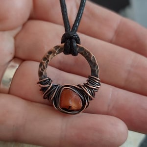May include: A copper wire wrapped pendant with a carnelian stone and a hammered copper circle. The pendant is attached to a black cord.