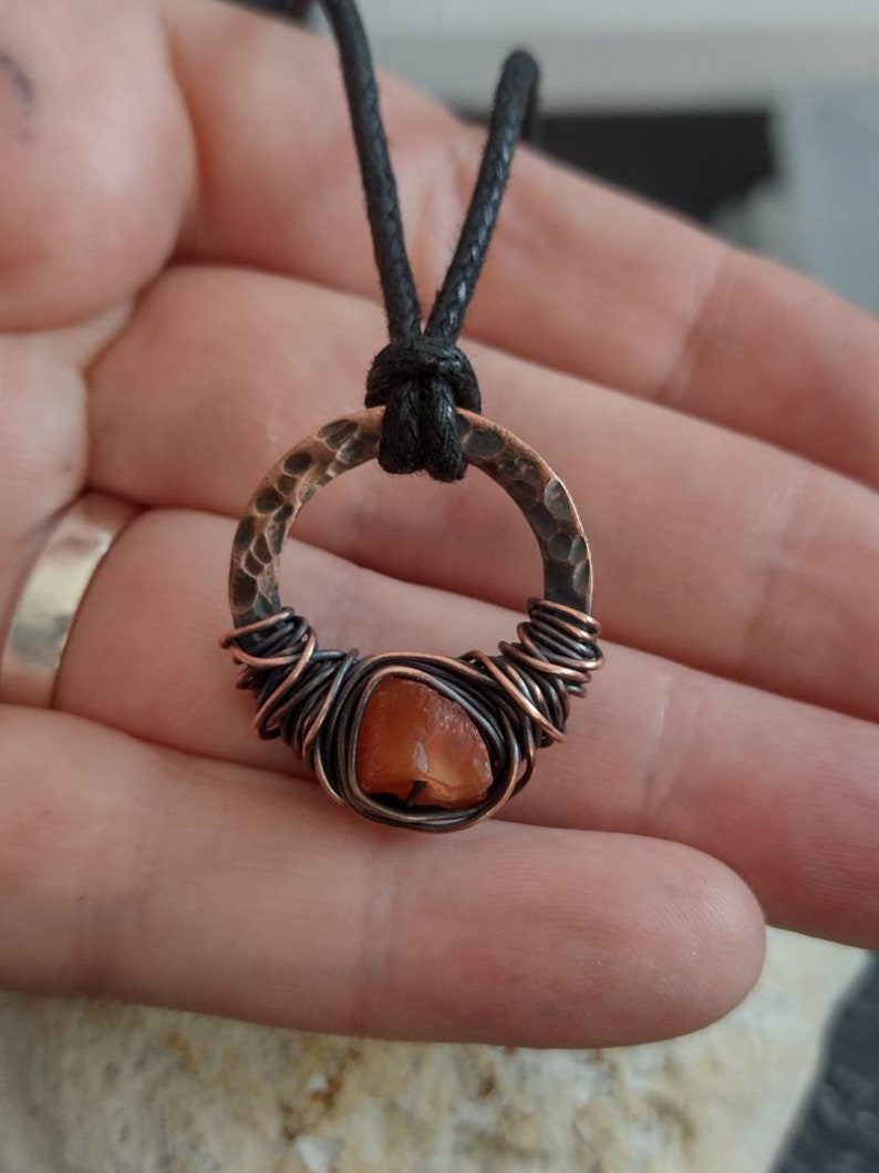 May include: A copper wire wrapped pendant with a carnelian stone and a hammered copper circle. The pendant is attached to a black cord.