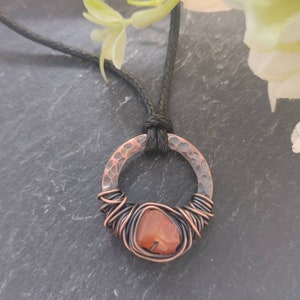 May include: A copper wire wrapped carnelian stone pendant necklace with a hammered copper circle. The necklace is on a black cord.