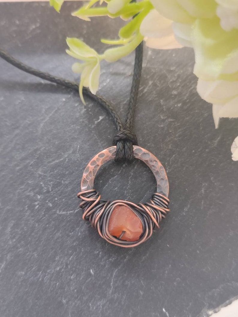 May include: A copper wire wrapped carnelian stone pendant necklace with a hammered copper circle. The necklace is on a black cord.