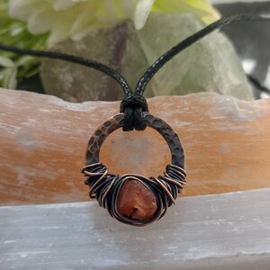 May include: A copper wire wrapped carnelian gemstone pendant on a black cord necklace. The pendant is a hammered copper circle with a carnelian stone in the center.