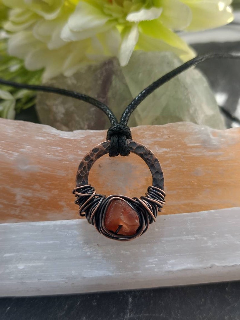 May include: A copper wire wrapped carnelian gemstone pendant on a black cord necklace. The pendant is a hammered copper circle with a carnelian stone in the center.