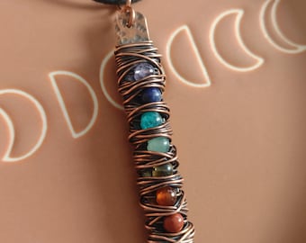 May include: A handmade pendant necklace featuring a copper-colored metal bar wrapped with wire and colorful beads. The beads are in shades of blue, green, orange, and brown. The pendant hangs from a black cord.