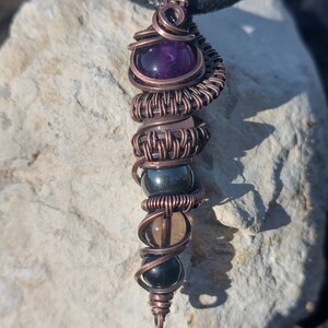 Op de afbeelding: Een hanger omwikkeld met koperdraad met een paarse amethiststeen, een heldere kwartsteen, een zwarte hematiet steen en een zwarte onyx steen.