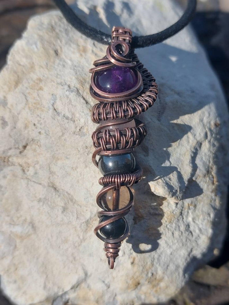 Op de afbeelding: Een hanger omwikkeld met koperdraad met een paarse amethiststeen, een heldere kwartsteen, een zwarte hematiet steen en een zwarte onyx steen.