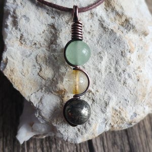 May include: A pendant featuring three stacked spherical beads in light green, translucent yellow, and metallic grey, secured by a coiled copper wire frame. The pendant hangs from a thin brown cord, showcasing a vertical arrangement of stones.