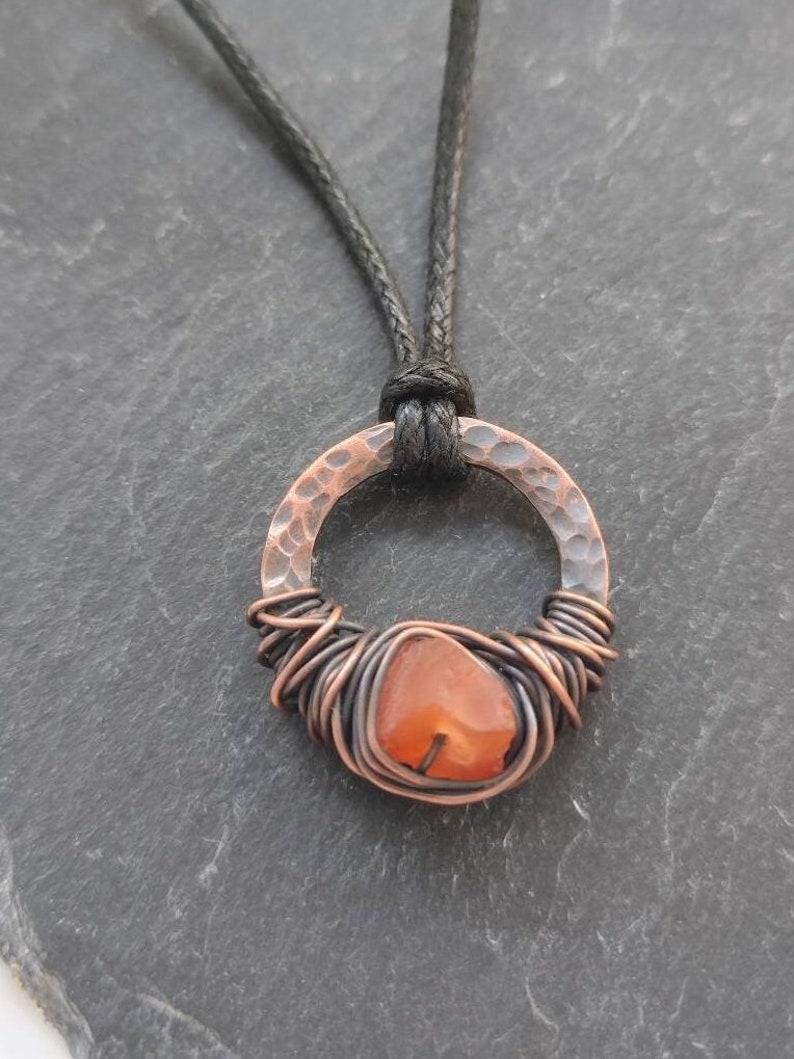 May include: A copper wire wrapped carnelian gemstone pendant with a hammered copper circle and a black cord.